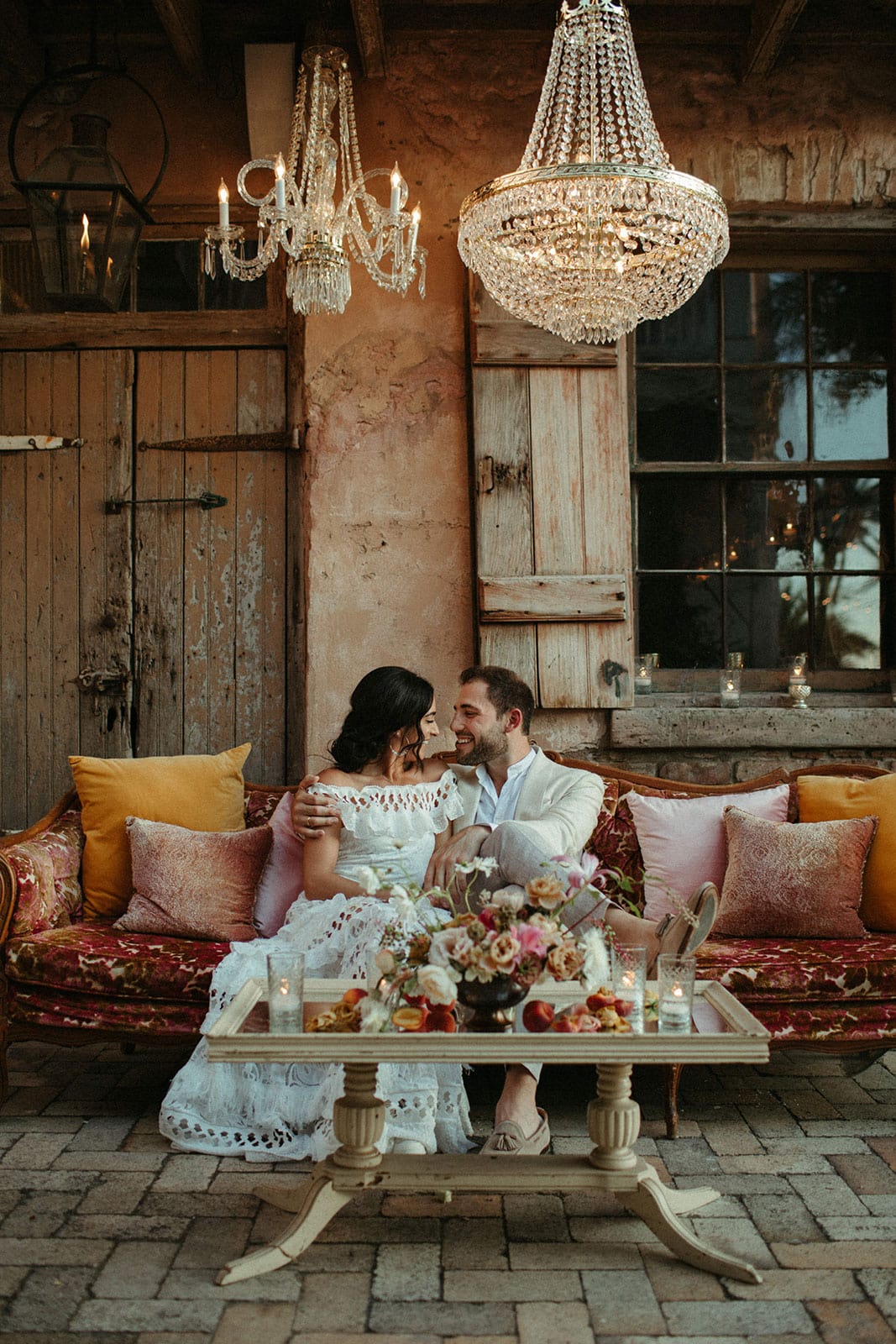 Brunette bride wearing Grace Loves Lace Coco Gown being held by groom sitting down on red sofa