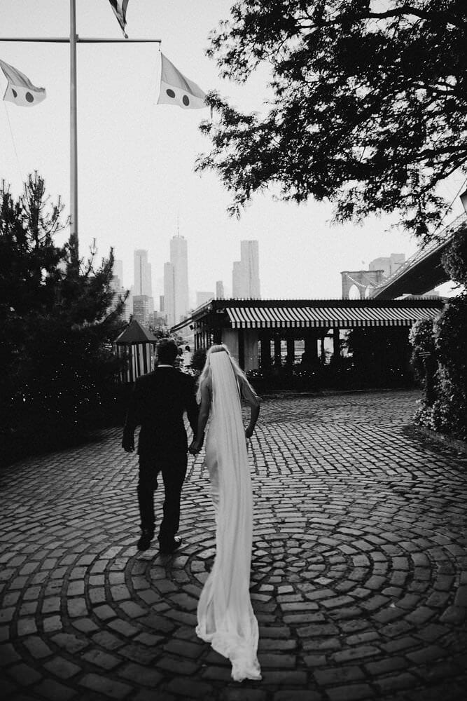 Black and white shot of bride wearing Grace Loves Lace Arlo Gown holding hands with groom in empty plaza