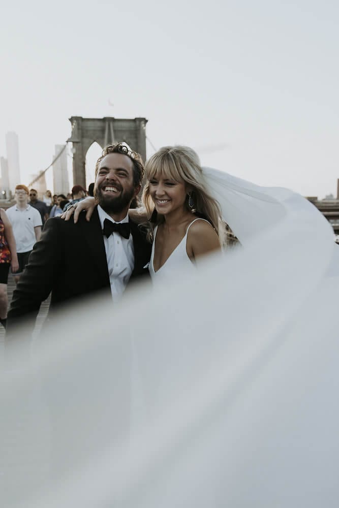 Blonde bride wearing Grace Loves Lace Arlo Gown embracing groom laughing with veil blowing in wind
