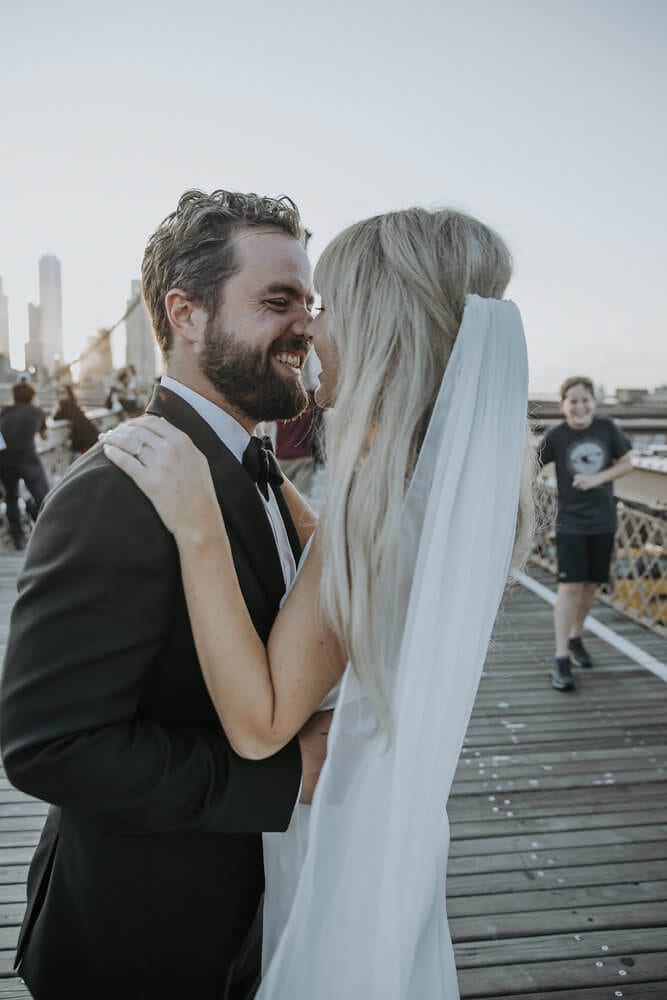 Blonde bride wearing Grace Loves Lace Arlo Gown embracing groom touching noses on bridge