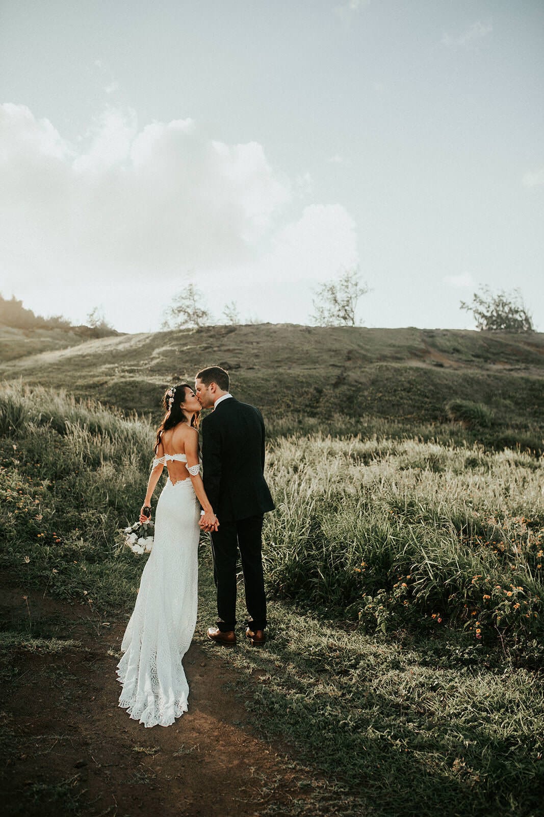 Brunette bride wearing Grace Loves Lace Paloma Gown kissing groom on green grassy hill