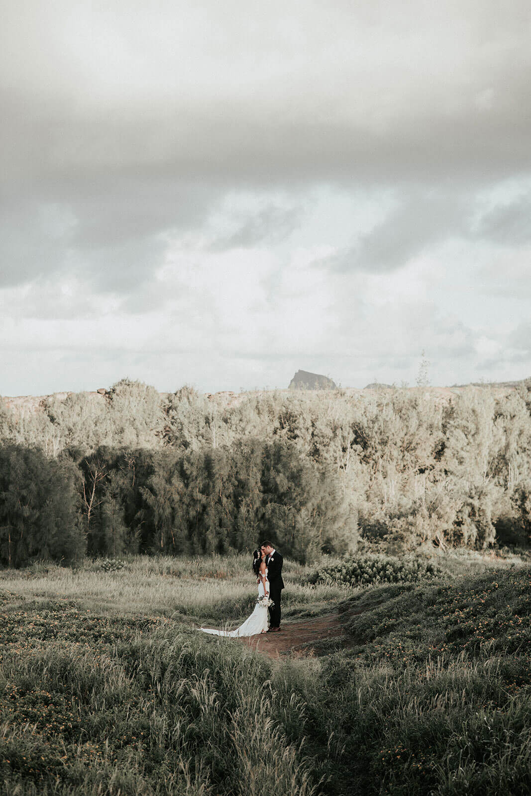 Brunette bride wearing Grace Loves Lace Paloma Gown kissing groom holding bouquet on grassy hill