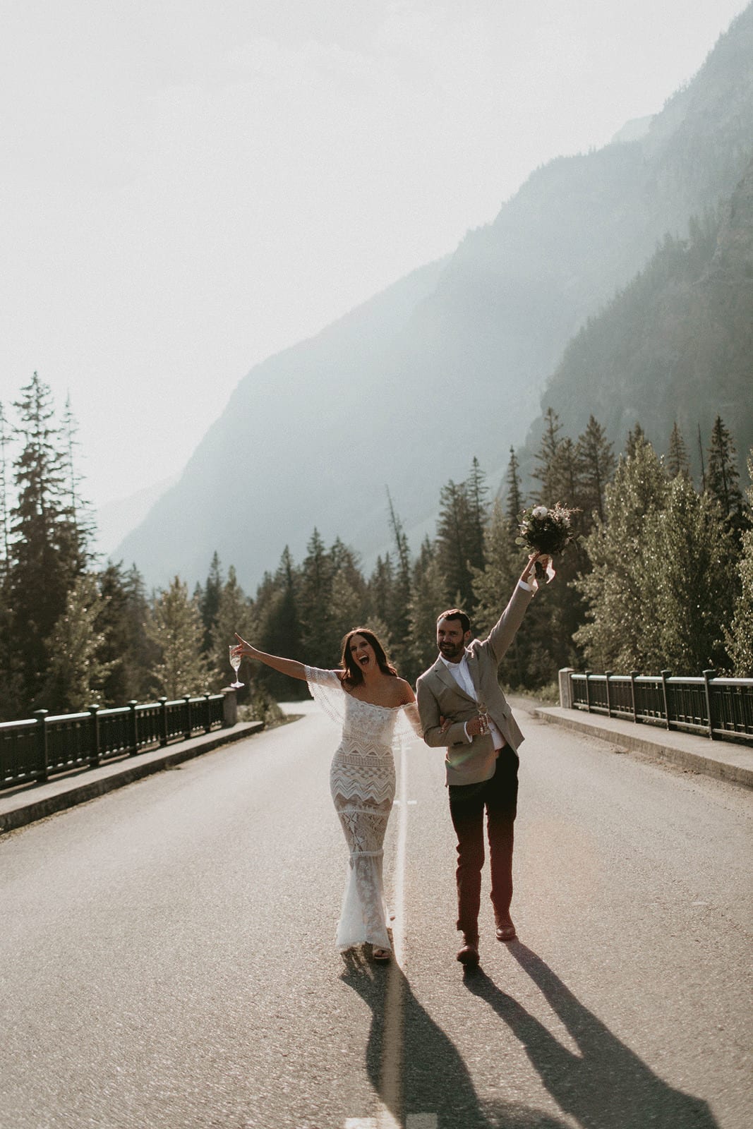 Brunette bride wearing Grace Loves Lace Emanuela Gown walking with groom on road with arms up
