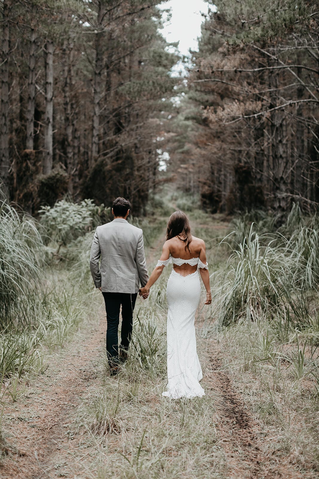 Brunette bride wearing Grace Loves Lace Paloma Gown holding hands with groom on forest path