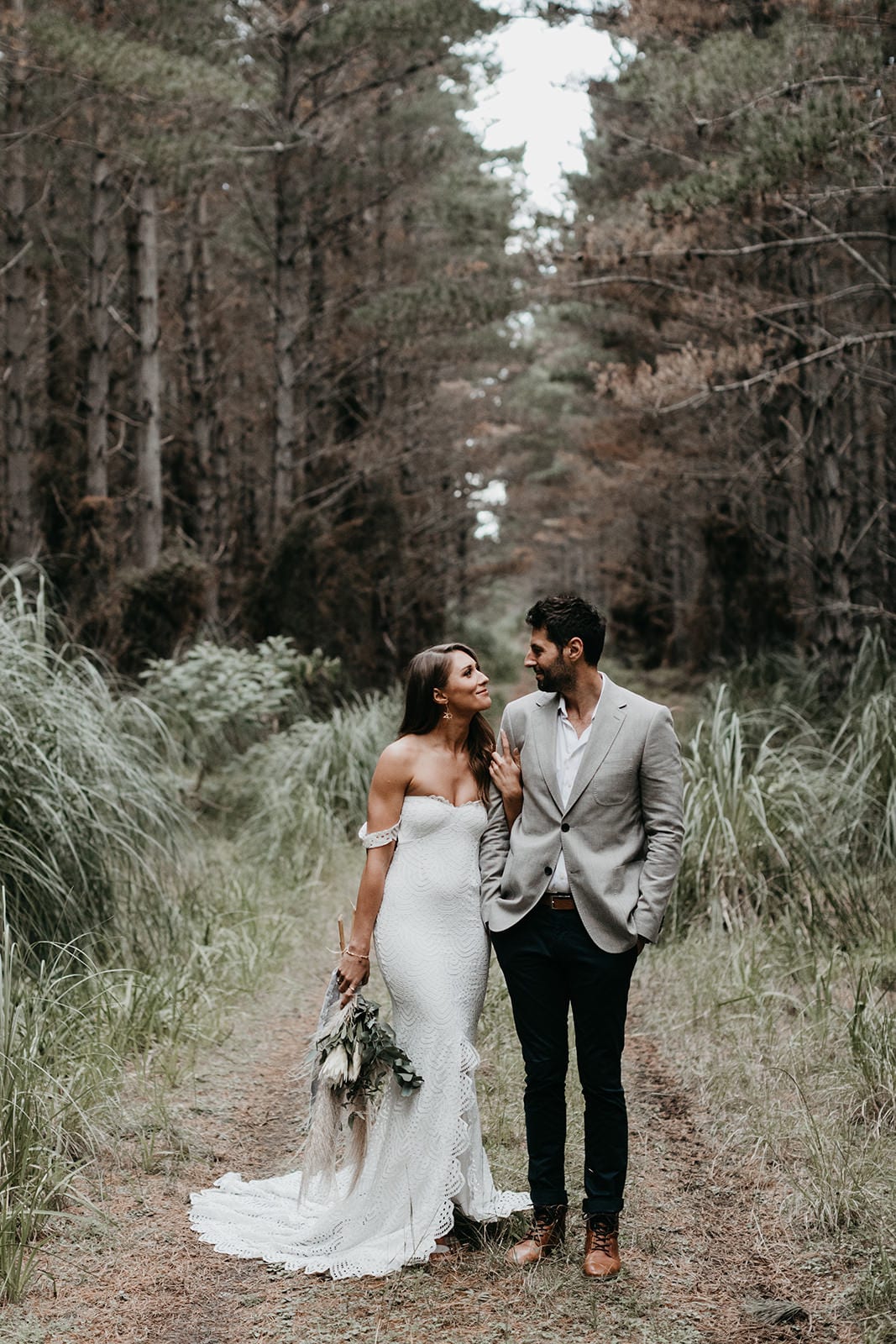 Brunette bride wearing Grace Loves Lace Paloma Gown looking at groom on forest path