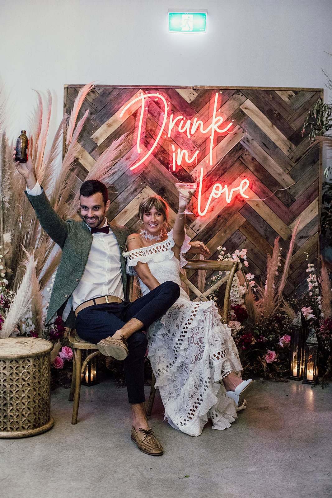 Blonde bride wearing Grace Loves Lace Coco Gown sitting with groom toasting in front of neon sign