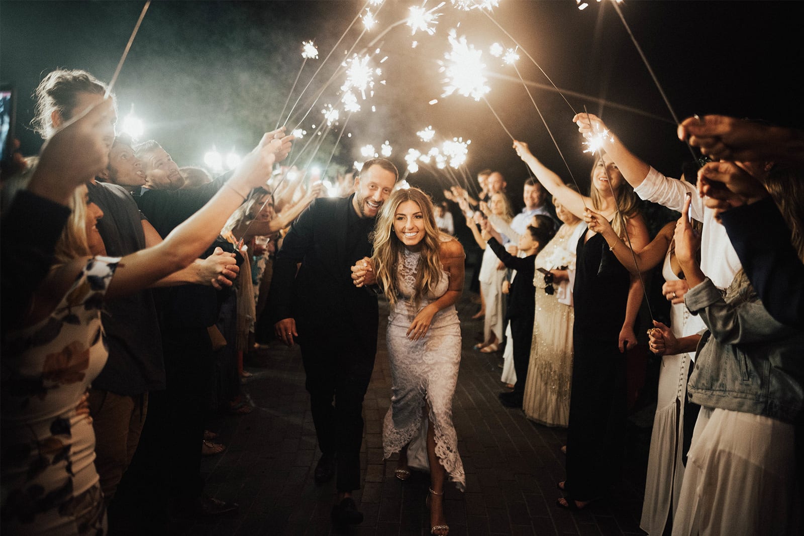 Bride wearing Grace Loves Lace Alexandra Gown holding hands with groom under archway made of sparklers